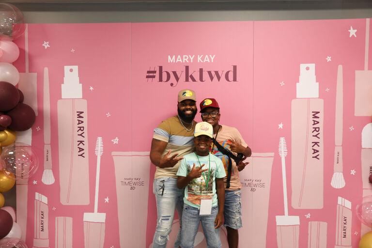 Tres personas posando frente a un fondo rosa de Mary Kay con gr&aacute;ficos de la marca y globos.
