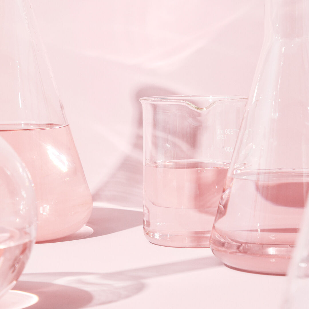 Recipientes de vidrio con l&iacute;quido transparente en una superficie rosa, reflejando ligeramente la luz rosa.