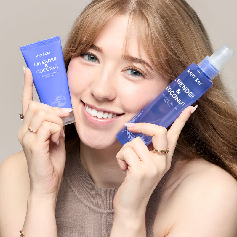 A white model with blue eyes holds the lavender and coconut hand cream and body spray by her face while smiling.&nbsp;