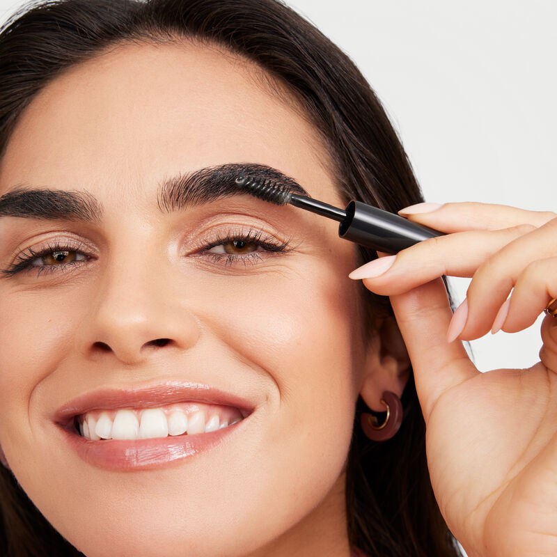 Smiling brunette model applying Mary Kay Clear Brow Styling Gel with applicator brush