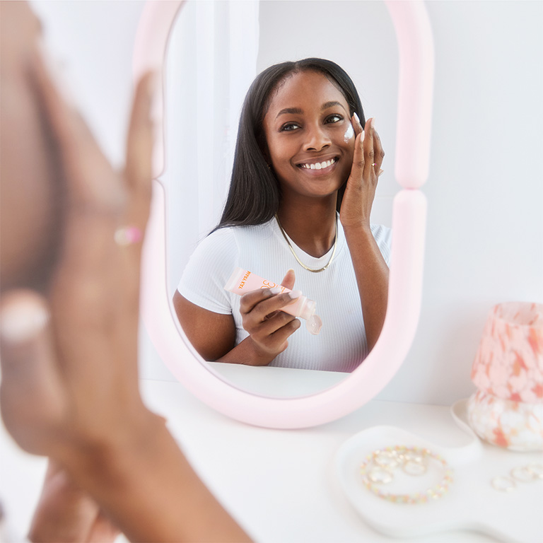 Mujer mir&aacute;ndose en un espejo de maquillaje y aplicando el Protector Solar Facial Mineral con FPS 30 Mary Kay&reg; en la mejilla.